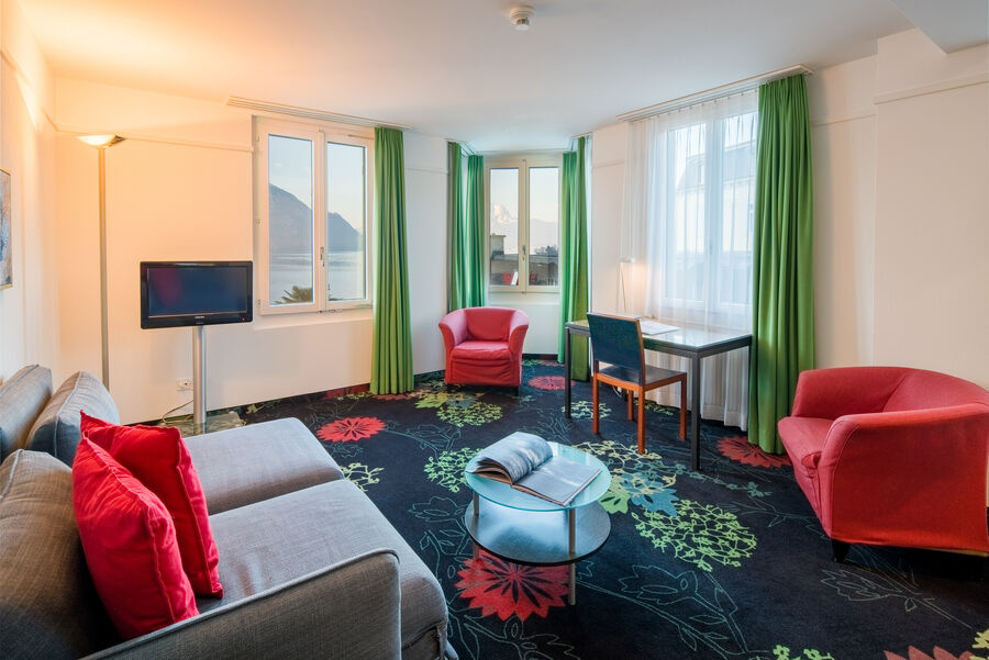 Junior Suite mit modernem Teppichboden, roten Sofas und einer grauen Couch. Zwei Fenster mit Blick auf den See