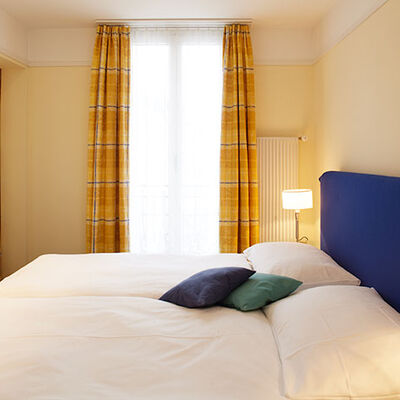 Gemütliches Budget-Zimmer mit grossem Bett und orange-gelben Gardinen. Zwei kleine Nachtlampen neben dem Bett